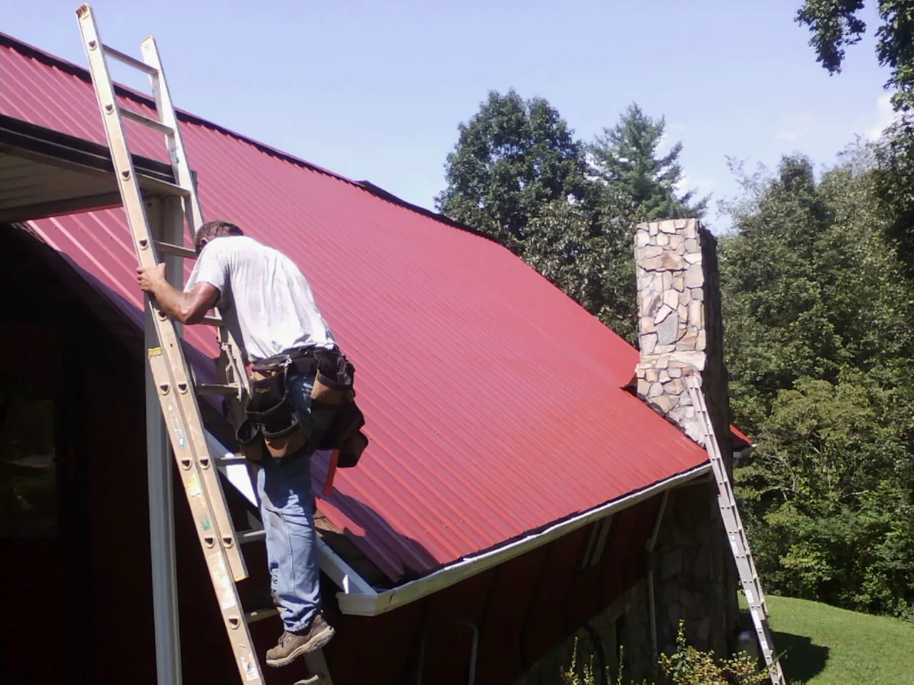 Roofer on a ladder beside a red metal roof and stone chimney — Soffit & Fascia Repair by Roofing Brothers in Faribault